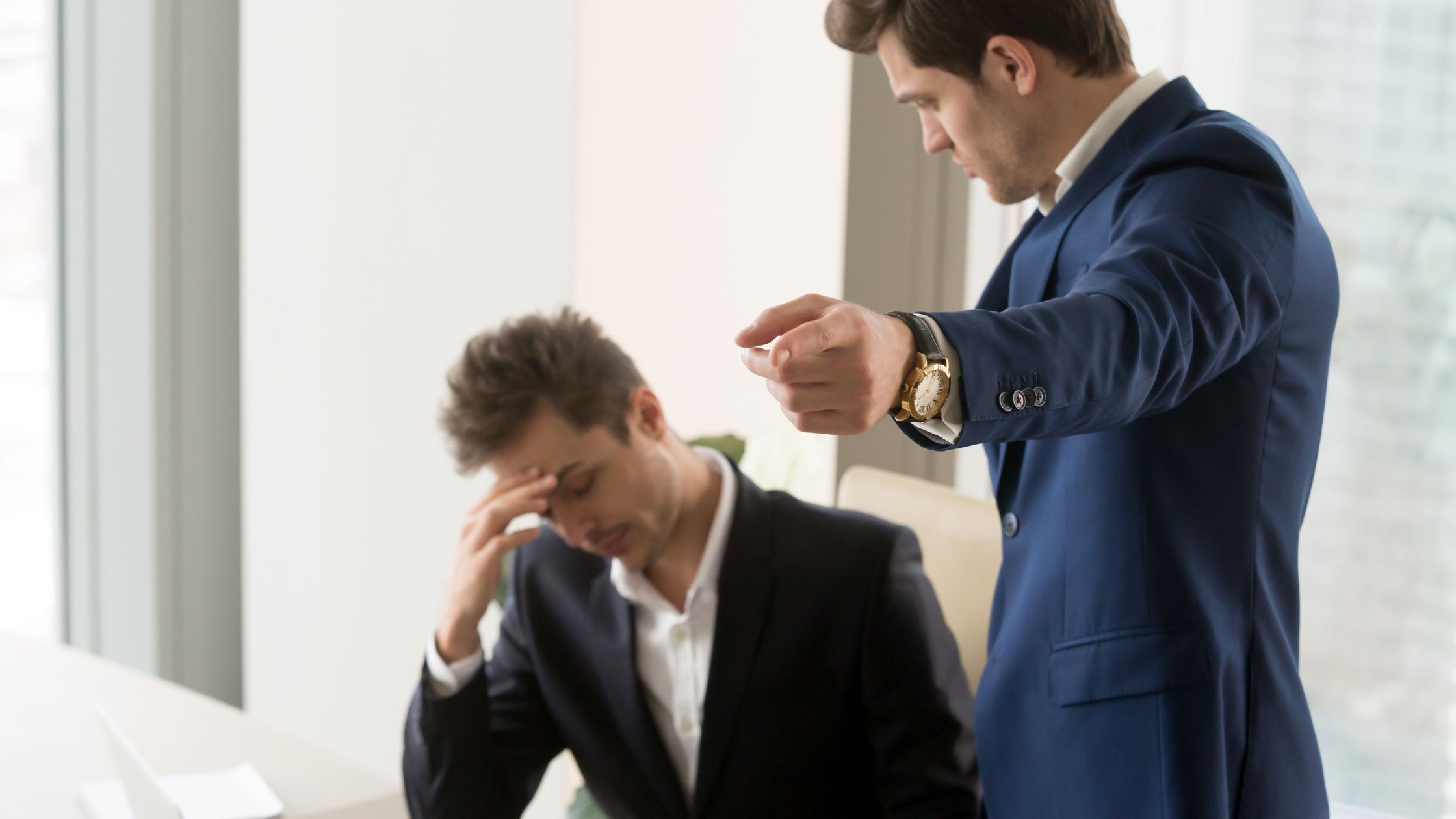 man in a business suit is pointing while another man in a suit sits with his head in his hand, appearing distressed. This image visually represents the challenging decision of firing a personal injury lawyer.